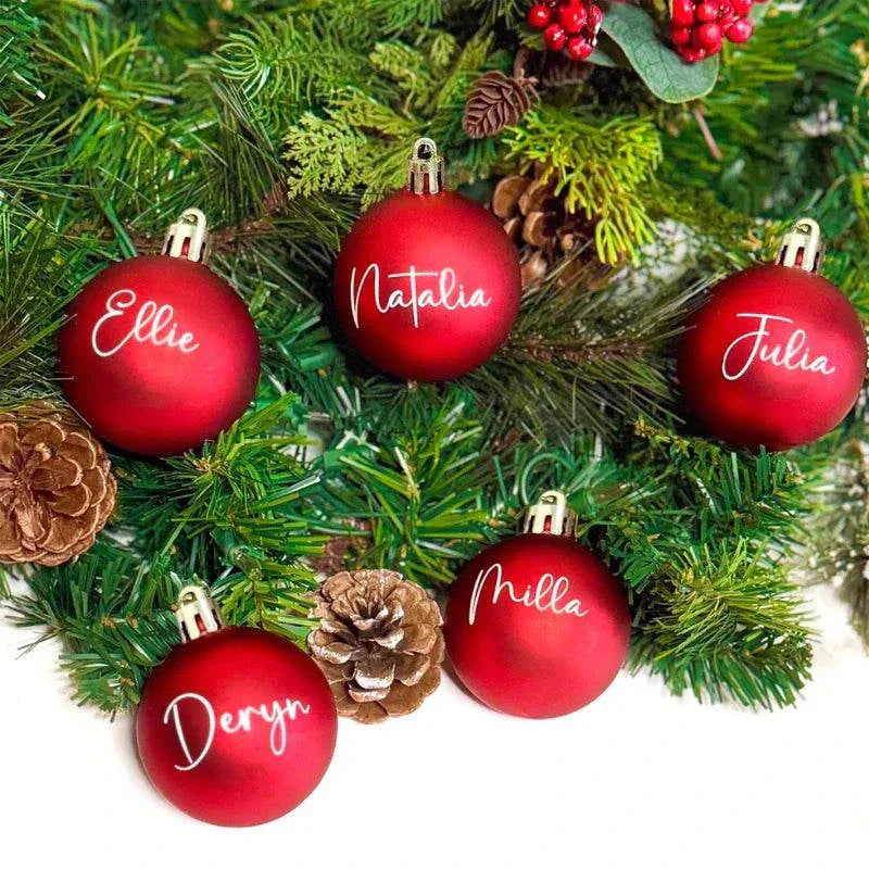 Personalized red Christmas balls with custom names hanging on green pine tree branches and pine cones